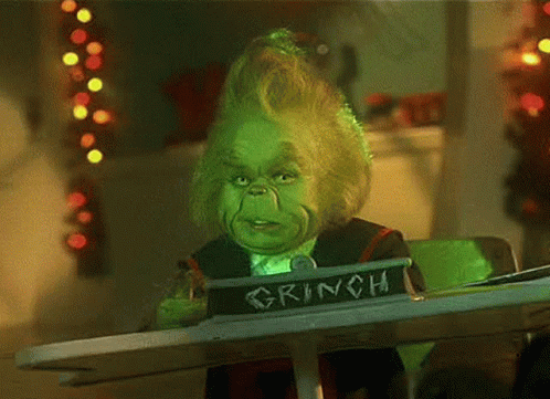 A Grinch Is Sitting At A Table With A Sign That Says Grinch On It . GIF