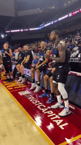 A Group Of Basketball Players Are Sitting On The Sidelines In Front Of A Sign That Says " Trust Capital " GIF