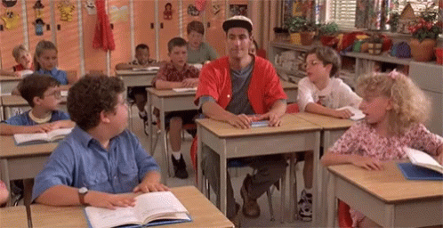 A Group Of Children Are Sitting At Their Desks In A Classroom With A Man In A Red Shirt Standing In The Middle . GIF