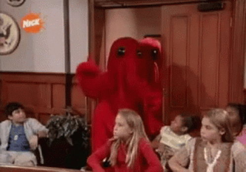 A Group Of Children Are Sitting In A Room With A Red Monster Mascot . GIF