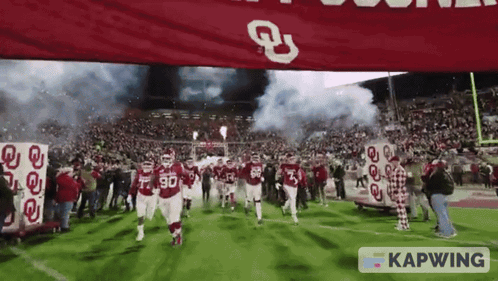 A Group Of Football Players Are Running On A Field With A Banner That Says Kapwing In The Foreground GIF