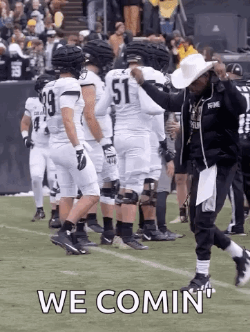 A Group Of Football Players Are Standing On A Field And A Man In A Cowboy Hat Is Walking On The Field . GIF