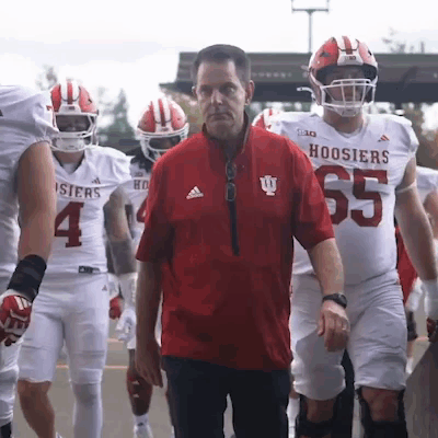 A Group Of Hoosier Football Players Are Walking Towards A Man In A Red Jacket GIF