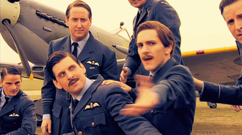 A Group Of Men Are Posing For A Picture In Front Of An Airplane And One Of Them Has The Letters Rnn On His Chest GIF