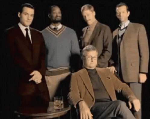 A Group Of Men Are Posing For A Picture With One Man Sitting In Front Of A Table With A Glass Of Whiskey GIF