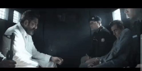 A Group Of Men Are Sitting In A Dark Room Talking To Each Other . One Of The Men Is Holding A Gun . GIF