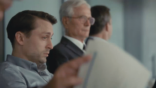 A Group Of Men Are Sitting In A Room And One Of Them Is Looking At A Piece Of Paper GIF