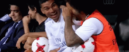 a group of men are sitting in a stadium with soccer balls and one of them is wearing a fly emirates shirt .