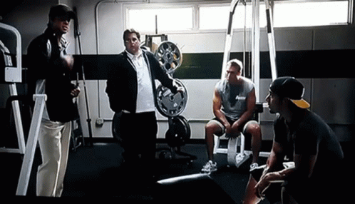 A Group Of Men Are Standing And Sitting In A Gym Talking To Each Other GIF
