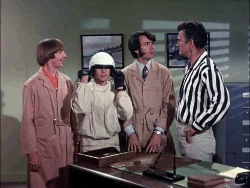 A Group Of Men Are Standing Around A Desk And One Of Them Is Wearing A White Helmet GIF