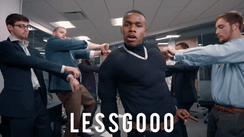 A Group Of Men Are Standing Around A Man In A Black Shirt With The Words Lessgood Written On It GIF