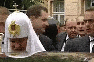 A Group Of Men Are Standing Around A Man Wearing A White Hat With A Cross On Top . GIF