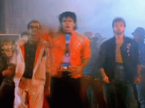 A Group Of Men Are Standing Next To Each Other In A Dark Room . One Of The Men Is Wearing An Orange Jacket . GIF