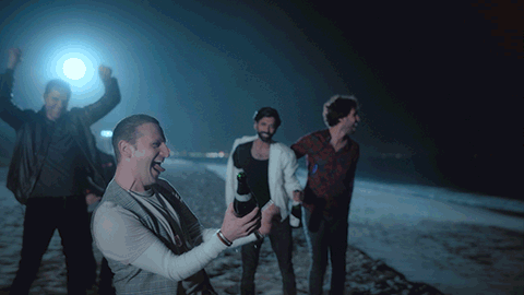 A Group Of Men Are Standing On A Beach At Night And One Of Them Is Holding A Bottle Of Beer GIF