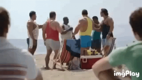 A Group Of Men Are Standing On A Beach With A Red Cooler . GIF