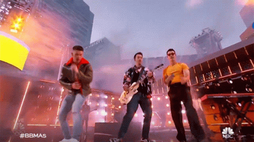 A Group Of Men Are Standing On A Stage Playing Instruments And Singing Into Microphones . GIF