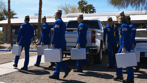 A Group Of Men In Blue Jumpsuits Are Carrying Briefcases In Front Of A Silver Truck That Says Gmc On The Back GIF