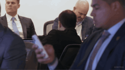 A Group Of Men In Suits And Ties Are Sitting Around A Table And One Man Is Holding A Cell Phone In His Hand GIF