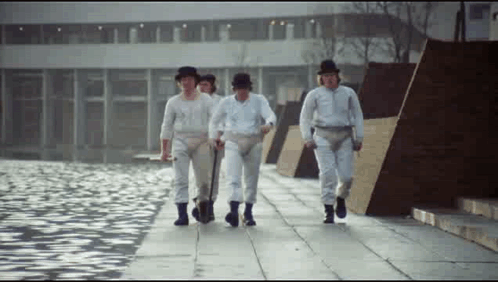 A Group Of Men In White Pants And Hats Are Walking Down A Sidewalk Near A Body Of Water . GIF