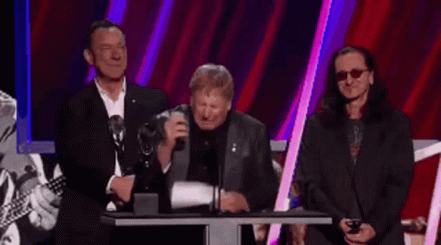 A Group Of Men Standing Around A Podium With One Wearing Glasses GIF