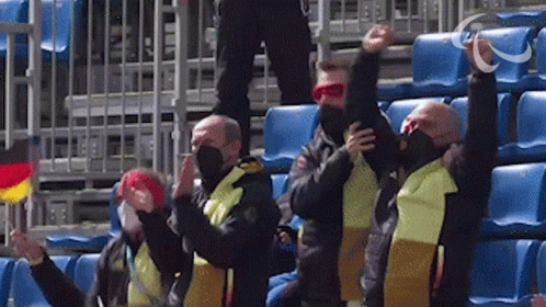 A Group Of Men Wearing Masks And Scarves Are Sitting In A Stadium With Their Arms In The Air GIF