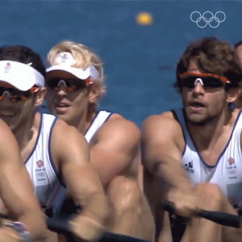 A Group Of Men Wearing Sunglasses And Visors Are Rowing In The Water With The Olympic Rings In The Background GIF