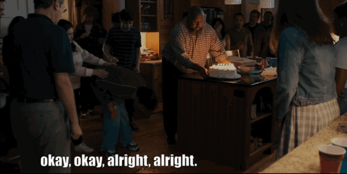 A Group Of People Are Gathered Around A Man Holding A Cake With The Words Okay Okay Alright Alright Below Him GIF