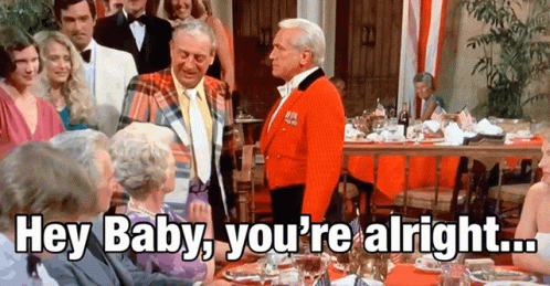 A Group Of People Are Gathered Around A Table With A Man In A Red Jacket Saying Hey Baby You 'Re Alright GIF