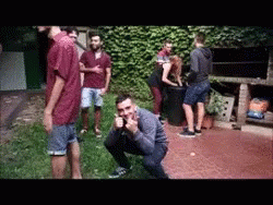 A Group Of People Are Gathered In A Backyard With One Man Squatting Down GIF