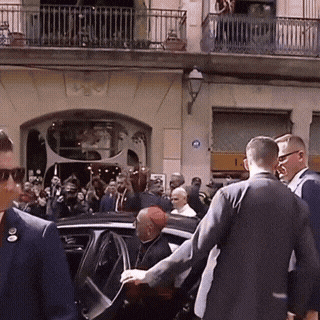 A Group Of People Are Gathered Outside A Building With A Blue Sign On The Wall That Says ' A ' On It GIF