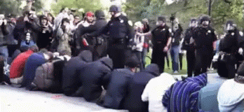 A Group Of People Are Kneeling Down In Front Of A Police Line GIF