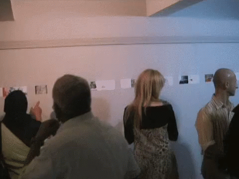 A Group Of People Are Looking At Pictures On A Wall And One Woman Is Pointing At One Of Them GIF