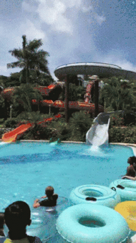A Group Of People Are Playing In A Swimming Pool With A Water Slide In The Background GIF