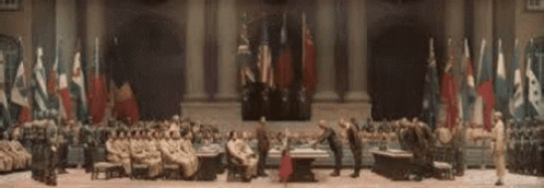 A Group Of People Are Sitting Around A Table In A Large Room With Flags Behind Them . GIF
