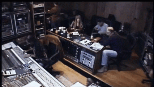 A Group Of People Are Sitting Around A Table In A Recording Studio With A Sign On The Wall That Says ' A ' GIF