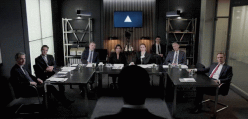 A Group Of People Are Sitting Around A Table With A Large Screen Behind Them That Says ' Triangle ' GIF
