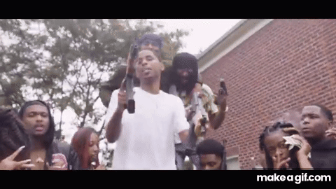 A Group Of People Are Standing Around A Man Holding A Gun In Front Of A Brick Building . GIF