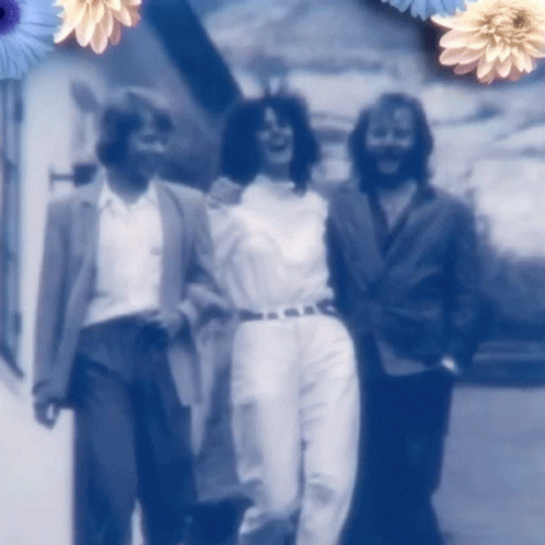 A Group Of People Are Walking Down A Street With Blue Flowers In The Background GIF