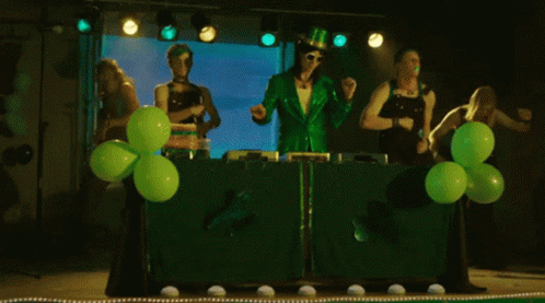 A Group Of People Dressed In Green Are Dancing In Front Of Green Balloons On A Stage GIF