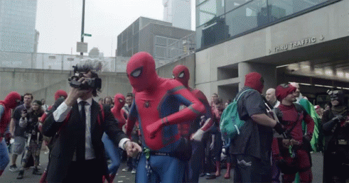 A Group Of People Dressed In Spiderman Costumes Are Walking Down A Street In Front Of A Building That Says Thru Traffic GIF