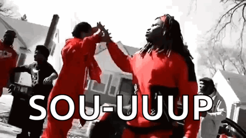 A Group Of People In Red Jumpsuits Are Dancing In Front Of A Building And A Sign That Says Sou-Uuup . GIF