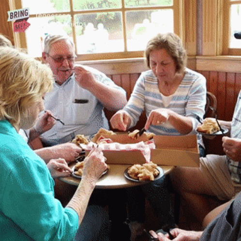 A Group Of People Sit Around A Table Eating Food With A Sign That Says Bring Me GIF