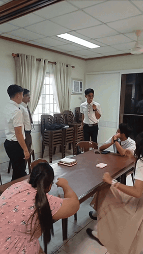 A Group Of People Sitting Around A Table With A Man Standing Behind Them Talking GIF