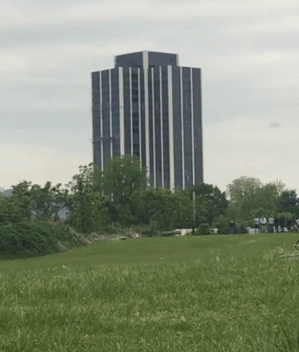 A Group Of People Standing In A Grassy Field With Smoke Coming Out Of A Building In The Background GIF