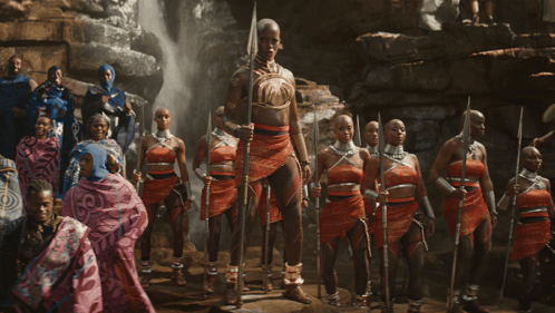 A Group Of People Standing In Front Of A Waterfall With One Woman Holding A Spear GIF