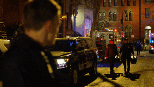 A Group Of People Walking Down A Street At Night With An Ambulance Behind Them GIF