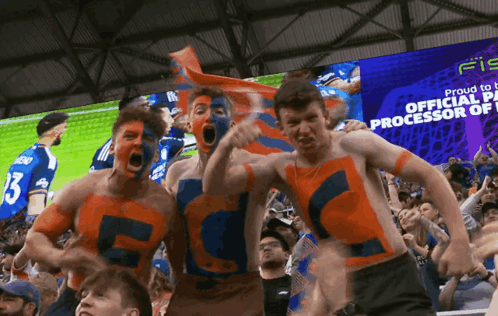 A Group Of Soccer Fans Are Standing In Front Of A Large Screen That Says Proud To Be Official Processor Of GIF