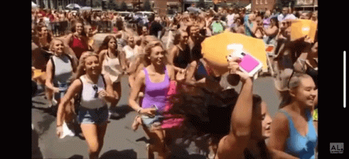A Group Of Women Are Running Down A Street With A Man Holding A Pillow That Says Al On It GIF