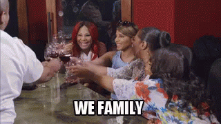 A Group Of Women Are Toasting With Wine Glasses At A Table With The Words `` We Family '' . GIF