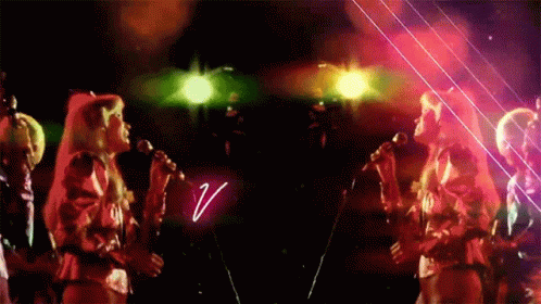 A Group Of Women Singing In Front Of A Neon Sign That Says V. GIF